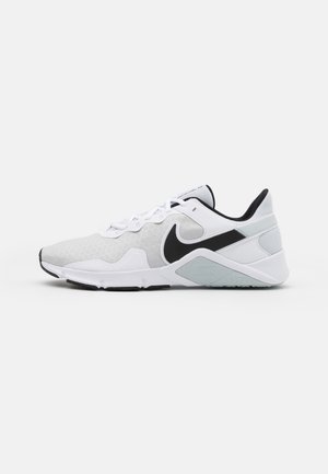 Biały i jasno szary sportowy but z czarnym logo Nike, białymi sznurowadłami i niskim kołnierzem przy kostce na jednolitym tle.