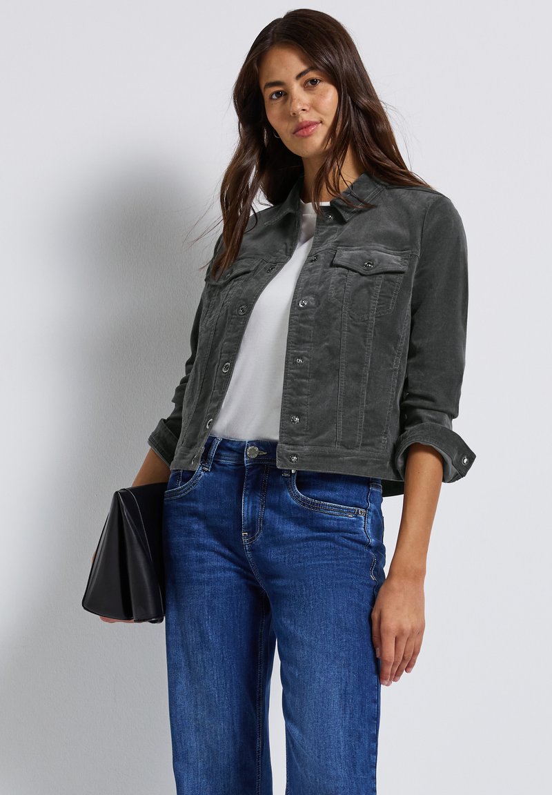 Šedá semišová bunda s knoflíkovým zapínáním, dvěma náprsními kapsami, ohrnutými rukávy, nošená přes bílé tričko a v kombinaci s modrými džíny.