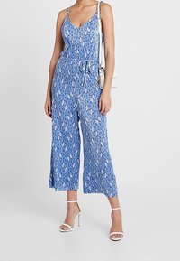 Modrý květinový overal s tenkými ramínky, výstřihem do V, nařaseným pasem a širokými kalhotami 3/4. Hladká tkanina a jemné vzorové detaily.