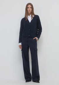 Marineblauer Cardigan mit fünf Knöpfen, über einem weißen Hemd mit Kragen getragen. Kombiniert mit marineblauen Cargohosen mit Nadelstreifen und seitlichen Taschen.