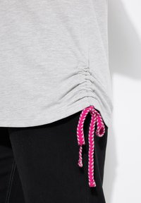 Top grigio a maniche corte con dettaglio arricciato sul lato e un accento con lacci rosa e bianchi all'orlo. Abbinato a pantaloni neri.