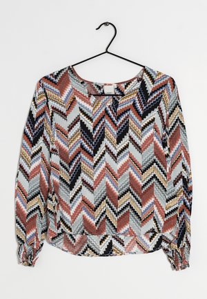 Dlouhý rukáv halenky s chevronovým cikcak vzorem v černé, bílé, rezavé, hořčicové a modré barvě, zavěšený na černém kovovém ramínku.