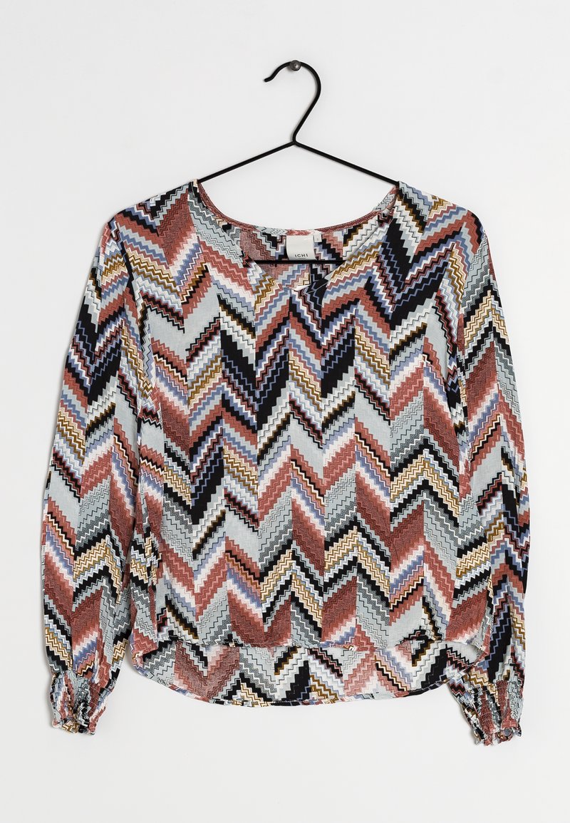 Blouse à manches longues avec un motif chevron en zigzag noir, blanc, rouille, moutarde et bleu, suspendue à un cintre en métal noir.