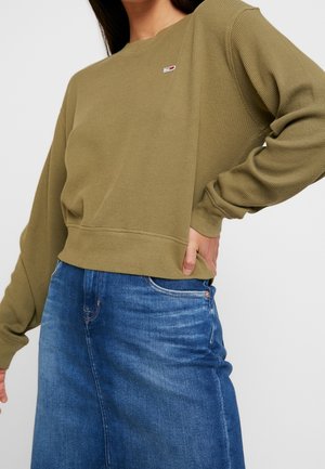 Vrouw met een olijfgroene, wafelgebreide trui met een klein logo en een blauwe denim rok, hand op de heup, tegen een effen achtergrond.