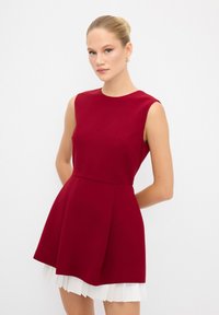 Robe rouge sans manches avec un corsage ajusté et une jupe évasée. Présente une doublure plissée blanche et une texture lisse et structurée.