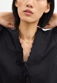 Blouse noire avec un décolleté festonné et un détail à nouer, accompagnée d'un collier en or avec un pendentif circulaire. Texture lisse, design minimaliste.