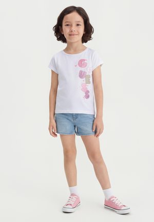 Pige med kort mørkt hår iført hvid Guess t-shirt, denimshorts, hvide sokker og lyserøde sneakers, stående mod en ensfarvet baggrund.