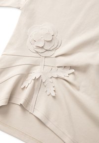 Beżowy t-shirt z teksturowanej tkaniny, z haftowanym wzorem kwiatu i liścia w podobnym odcieniu, tworzącym subtelny, warstwowy efekt.