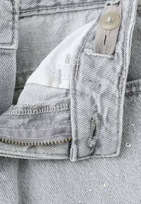 Šedá džínová látka se zipem, zapínáním na knoflíky a bílou podšívkou. Obsahuje malé lesklé detaily a viditelné stehy.