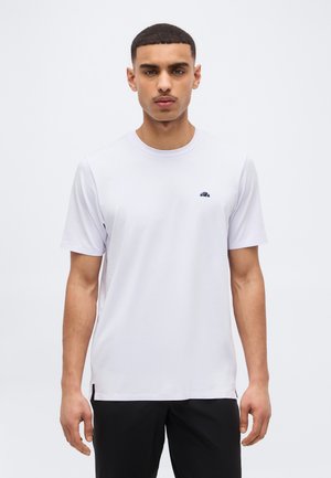 Jeune homme aux cheveux courts et foncés, portant un t-shirt blanc uni à manches courtes et un pantalon noir, debout devant un fond blanc.