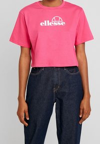 Camiseta corta rosa de algodón con cuello redondo y mangas cortas, que presenta un gráfico de logo "ellesse" en blanco en la parte frontal.
