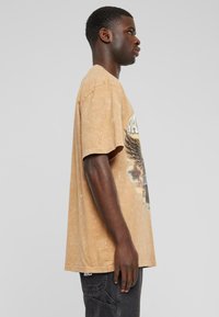 Tan oversized t-shirt med en falmet finish, prydet med en grafisk trykning af vinger og tekst på fronten. Kortærmet og med en afslappet pasform.