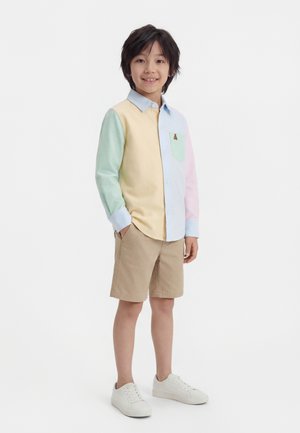 Παιδί με σκουρόχρωμα μαλλιά που φοράει πολύχρωμο πουκάμισο με μακριά μανίκια και κουμπιά, μπεζ σορτς και λευκά αθλητικά παπούτσια, στέκεται με τα χέρια στις τσέπες.