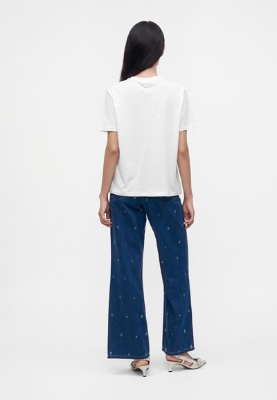 Femme aux longs cheveux foncés portant un t-shirt blanc uni à manches courtes, un jean large bleu avec de petits motifs blancs et des sandales blanches à talons.