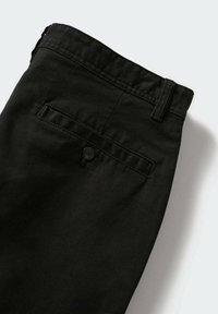 Pantalon noir montrant une poche arrière avec fermeture par bouton et passants pour ceinture sur un fond clair uni.