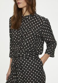 Schwarzes Hemd mit Knopfleiste und dreiviertel Ärmeln, mit einem floralen, kreisförmigen Muster in Weiß und Beige. Taillierter Knotenakzent. Weiche Stoffstruktur.