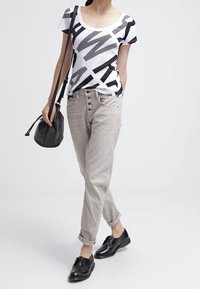 Tričko s černobílým vzorem, světle šedé volné kalhoty s detailním knoflíkem, černé kožené boty a malá černá crossbody taška.