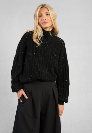 Donna bionda che indossa un maglione a collo alto nero a coste con paillettes e pantaloni larghi neri con tasca con bottone, in piedi con una mano nella tasca.