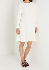 Langärmliges, cremefarbenes Kleid mit vertikaler Rippstruktur und lockerem Schnitt, kombiniert mit schwarzen Riemchensandalen.