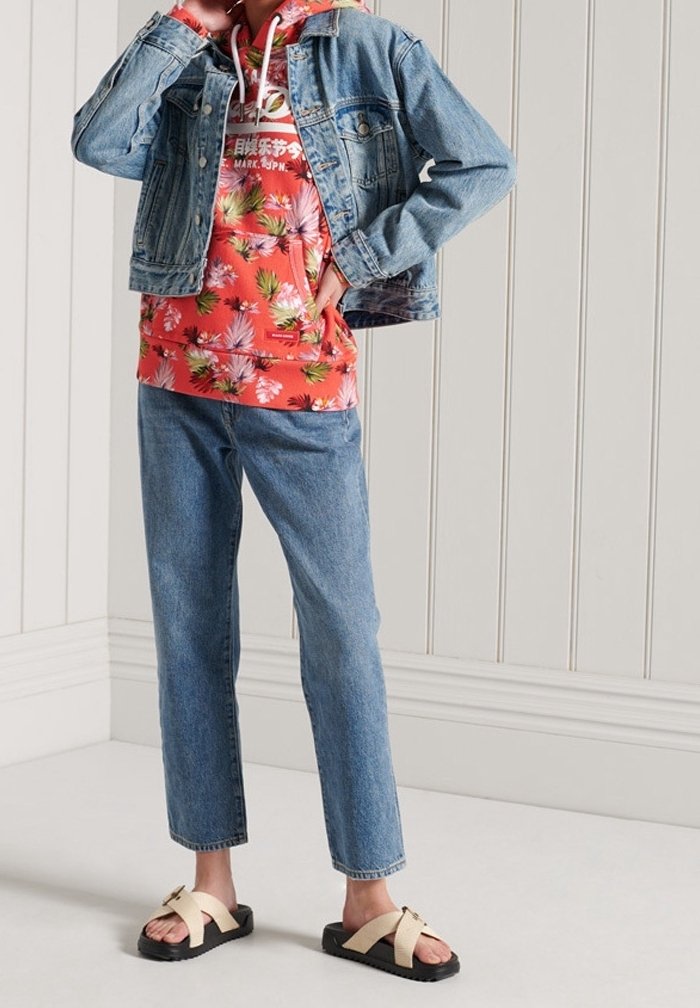 Personne portant une veste en jean par-dessus un sweat à capuche rouge à fleurs, un jean bleu et des sandales beiges à brides croisées, debout contre un mur blanc à panneaux.