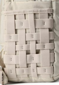 Mochila de tela tejida beige con un patrón de malla texturizada y tirantes similares a cintas, que presenta un material liso y duradero en el cuerpo.