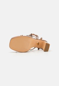 Beige høyhælte sandaler med en kraftig, teksturert såle og slanke ankelstropper. Har en rund hæl, glatt overflate og logo-detalj på siden.