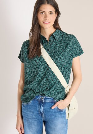 Frau steht vor einfarbigem Hintergrund, trägt ein grünes kurzärmeliges Hemd mit Muster, blaue Jeans und einen beigen Umhängegurt über der Schulter.