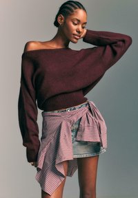 Bordo swetere z off-shoulder, izdelan iz mehkega materiala, v kombinaciji z našitkano krilo s črtami v rdeči in beli barvi, ki ga nosimo čez jeans kratke hlače.
