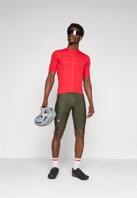 Maillot de cyclisme rouge à manches courtes avec une fermeture éclair à l'avant, shorts de cyclisme vert olive, chaussures de cyclisme noires, et un casque gris à la main.