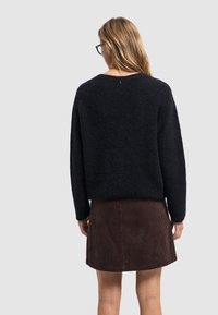 Zwarte, gestructureerde trui met wijde mouwen, gecombineerd met een bruine corduroy rok, van achter bekeken tegen een grijze achtergrond.