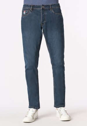 Jeans in denim blu scuro con vestibilità slim, cinque tasche e cuciture color beige. Abbinati a sneakers bianche per un look casual.