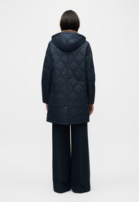 Κουκούλα σκούρου μπλε παλτό με καπιτονέ σχεδιασμό, με μήκος μέχρι τη μέση και ορατό μοτίβο ραφής. Φορεμένο πάνω από σκούρο παντελόνι με φαρδιά πόδια.