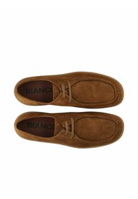 Zapatos de ante marrón con puntera redondeada, detalles de costura y cordones finos. El interior presenta un estampado de logo en negro.