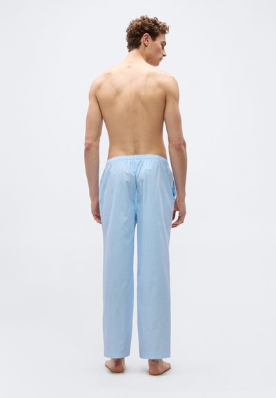 Νεαρός άνδρας στέκεται ξυπόλητος, χωρίς πουκάμισο, φορώντας ανοιχτό μπλε και λευκό παντελόνι πιτζάμας με κατακόρυφες ρίγες, με την πλάτη προς το μέρος μας σε απλό φόντο.