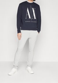 Felpa blu nave con logo bianco "AX" e testo "ARMANI EXCHANGE". Abbinata a pantaloni slim-fit grigi chiari e sneakers bianche. Tessuto liscio.
