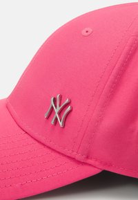 Rózsaszín baseballsapka, amely szövetből készült, ezüst hímzett "NY" logóval. Lekerekített napszemüveg és struktúrált korona, látható varrással.