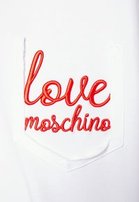 Vitt tyg med ett rött broderat mönster med texten "love moschino" på en ficka. Fickan har en rundad, diamantliknande form.