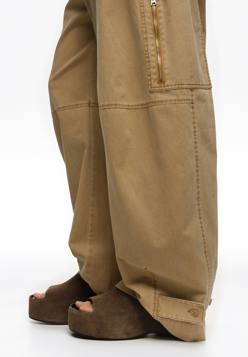 Pantalones de pierna ancha en color beige con un gran bolsillo frontal y detalle de cremallera, combinados con zapatos plataforma abiertos de ante marrón.