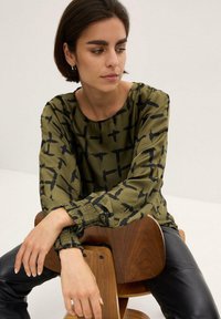 Blusa verde oliva con un motivo astratto nero, maniche lunghe e polsini arricciati. Abbinata a pantaloni in pelle ecologica neri.