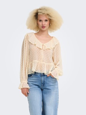 Femme aux cheveux bouclés blonds portant un chemisier transparent crème avec des volants et un jean taille haute bleu clair, debout devant un fond uni.