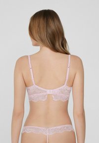 Soutien-gorge en dentelle rose tendre avec bretelles réglables, présentant des bords festonnés et une fermeture à crochets dans le dos. Design en dentelle détaillée sur l'ensemble.