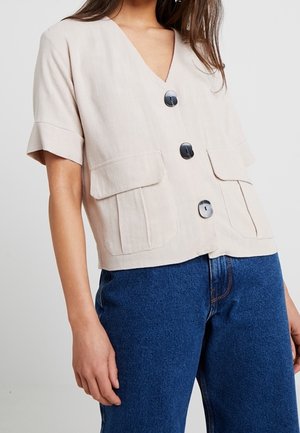 Kobieta w beżowej, krótkim rękawem, zapinanej na guziki bluzce z dużymi kieszeniami, zestawionej z niebieskimi dżinsami z wysokim stanem.