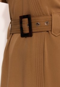 Brązowa sukienka z materiału z szerokim paskiem, posiadająca prostokątną klamrę w kolorze skorupy żółwia oraz zdobienia w postaci przeszyć po bokach.