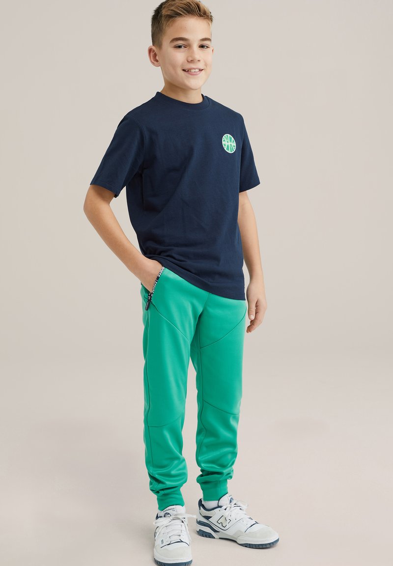 Navy cotton t-shirt with a round green basketball logo, paired with green tapered joggers featuring seams and zipper details.