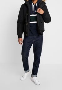 Sort pufferjakke med hætte foret med pels, båret over en navy sweatshirt med hvide og grønne striber, kombineret med mørk denim og hvide sneakers.