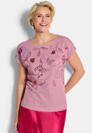 Lächelnde Frau mit kurzem blondem Haar, die ein rosa, mit floralen Stickereien verziertes Oberteil und einen leuchtend pinken Rock trägt, vor einem weißen Hintergrund stehend.