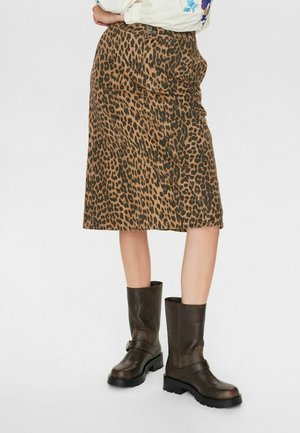 Femme portant une jupe marron à imprimé léopard longueur genou et des bottes en cuir marron foncé montant au milieu du mollet, debout sur fond blanc.