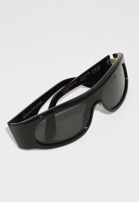 Black sunglasses with a sleek, rounded design. Made from glossy material, featuring dark lenses and gold branding on the temple.