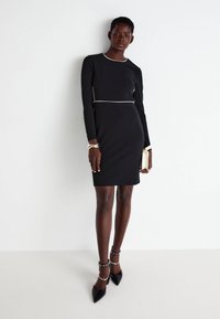 Μαύρο φόρεμα bodycon με μακριά μανίκια και λευκές πινελιές. Διαθέτει στρογγυλή λαιμόκοψη και στενή σιλουέτα, συνδυασμένο με μυτερές γόβες.