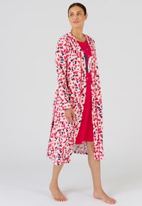 Patroon lange cardigan in lichtroze met rode en blauwe hartvormen, met lange mouwen en een eenvoudige open voorkant, gedragen over een rode jurk.